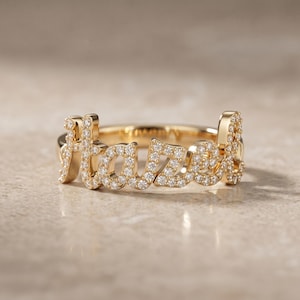 May include: A gold-colored ring with the name "Hazel" spelled out in sparkling, clear gemstones. The ring is set against a neutral background, highlighting the intricate design and the brilliance of the stones. The ring is a piece of jewelry.