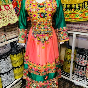 May include: A vibrant traditional dress with detailed embroidery. The bodice is embellished with gold and colourful accents, complemented by green sleeves and a peach skirt. The dress includes decorative mirrors and a detailed hemline, reflecting cultural artistry.