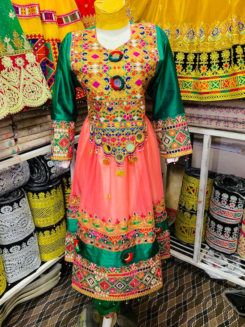 May include: A vibrant traditional dress with detailed embroidery. The bodice is embellished with gold and colourful accents, complemented by green sleeves and a peach skirt. The dress includes decorative mirrors and a detailed hemline, reflecting cultural artistry.