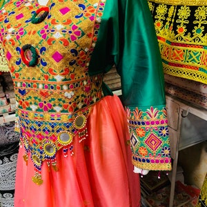 May include: A detailed traditional dress with colourful embroidery. The bodice is adorned with gold, pink, and blue patterns, complemented by green sleeves and a peach skirt. The waistline features mirrors and beaded embellishments.