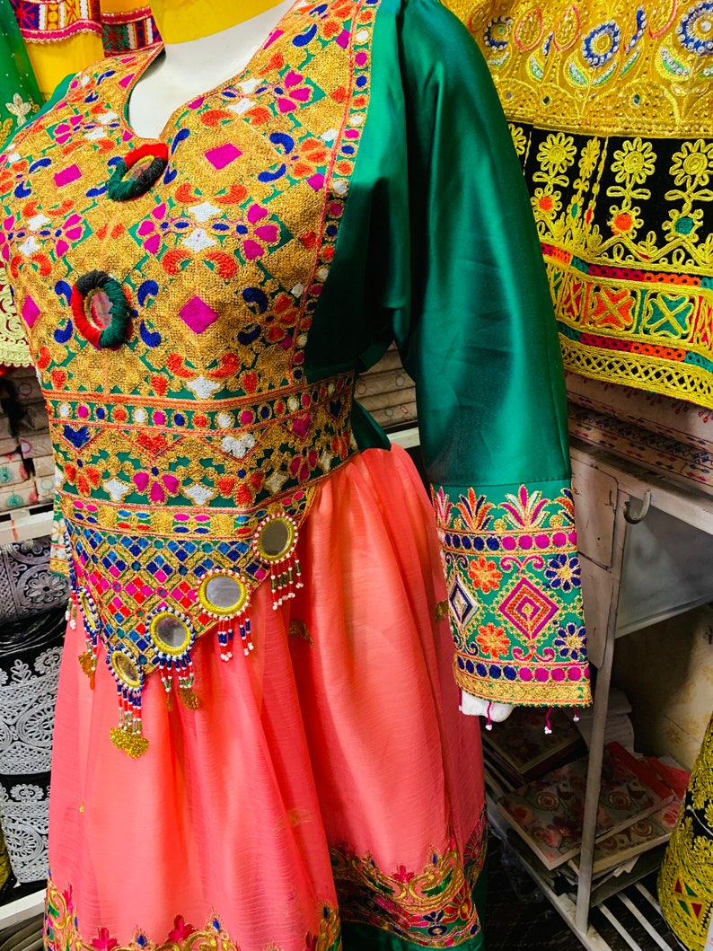 May include: A detailed traditional dress with colourful embroidery. The bodice is adorned with gold, pink, and blue patterns, complemented by green sleeves and a peach skirt. The waistline features mirrors and beaded embellishments.