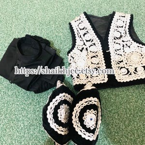 May include: A black outfit with a white embroidered vest and two black hats with white trim. The vest has a black base with white floral embroidery. The hats have a similar design, with a blue gem in the center. The outfit is displayed on a green surface.