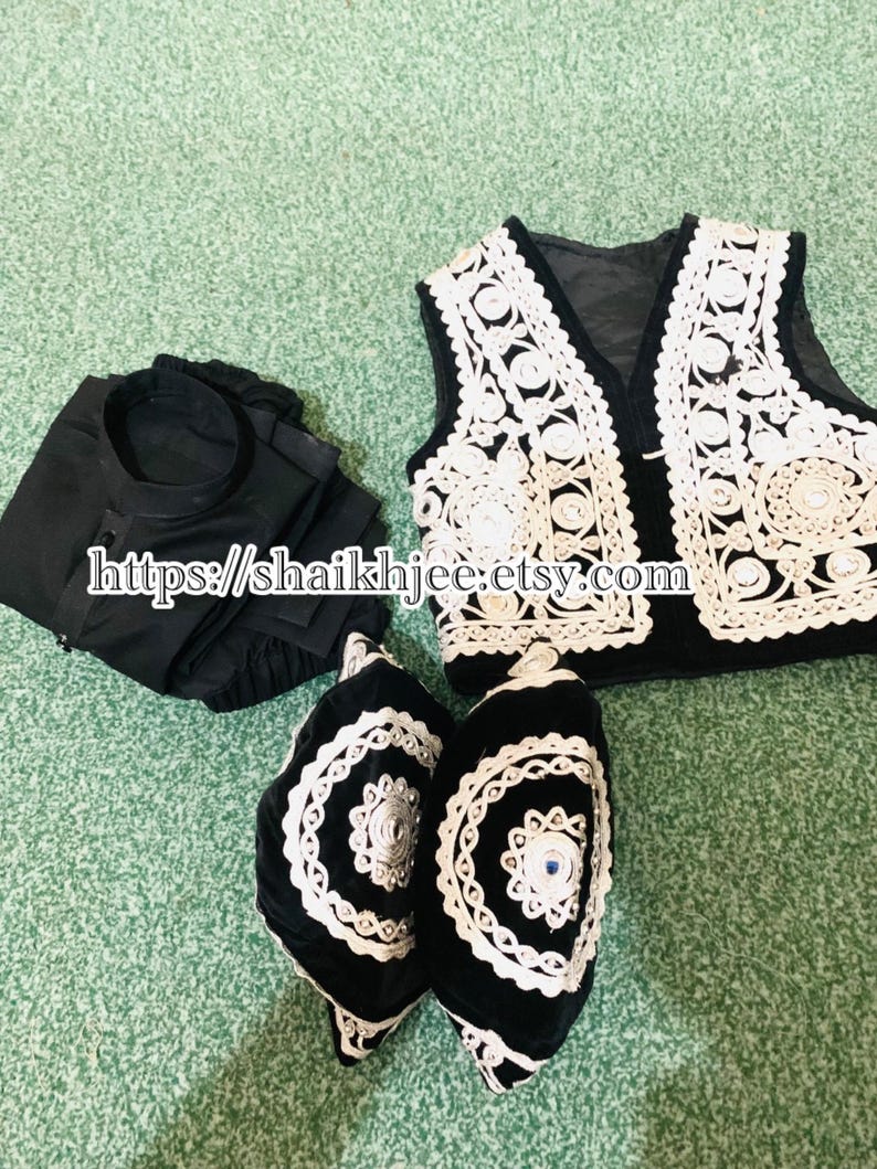 May include: A black outfit with a white embroidered vest and two black hats with white trim. The vest has a black base with white floral embroidery. The hats have a similar design, with a blue gem in the center. The outfit is displayed on a green surface.