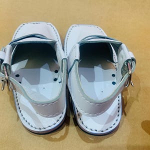 May include: A pair of white leather sandals with a strap and buckle closure. The sandals have a stitched design around the edges and a dark blue sole. The sandals are designed for children.