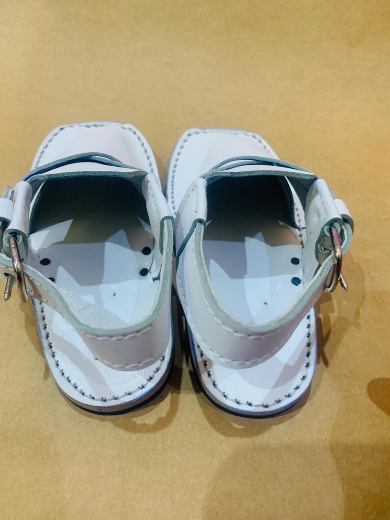 May include: A pair of white leather sandals with a strap and buckle closure. The sandals have a stitched design around the edges and a dark blue sole. The sandals are designed for children.