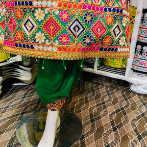 May include: A green garment with detailed embroidery in pink, blue, orange, and gold. The design incorporates floral and geometric patterns. The garment is displayed on a white mannequin leg. The embroidery is very detailed.