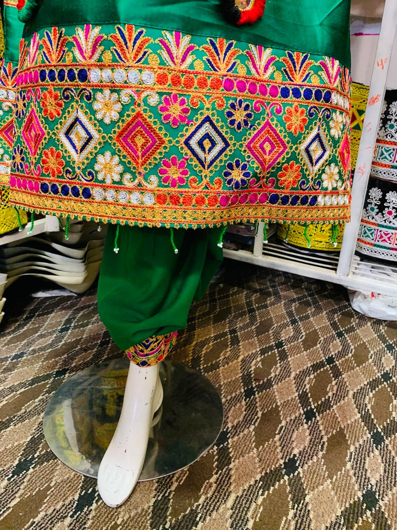 May include: A green garment with detailed embroidery in pink, blue, orange, and gold. The design incorporates floral and geometric patterns. The garment is displayed on a white mannequin leg. The embroidery is very detailed.