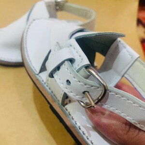 May include: White leather sandals with a silver buckle closure. The sandals have a stitched design around the edges and a light brown sole. The straps are adjustable for a comfortable fit. These sandals are perfect for warm weather.