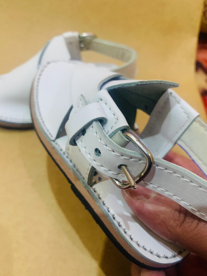 May include: White leather sandals with a silver buckle closure. The sandals have a stitched design around the edges and a light brown sole. The straps are adjustable for a comfortable fit. These sandals are perfect for warm weather.