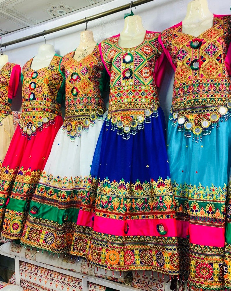 May include: A display of embroidered dresses in various colours, including red, white, blue, and green. The dresses are adorned with intricate patterns and mirror embellishments. They are hanging on a metal rail.