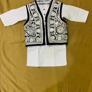 May include: A white kurta and matching pants set with a black vest. The vest is decorated with intricate silver embroidery and small reflective mirrors. The outfit is displayed on a tan surface.