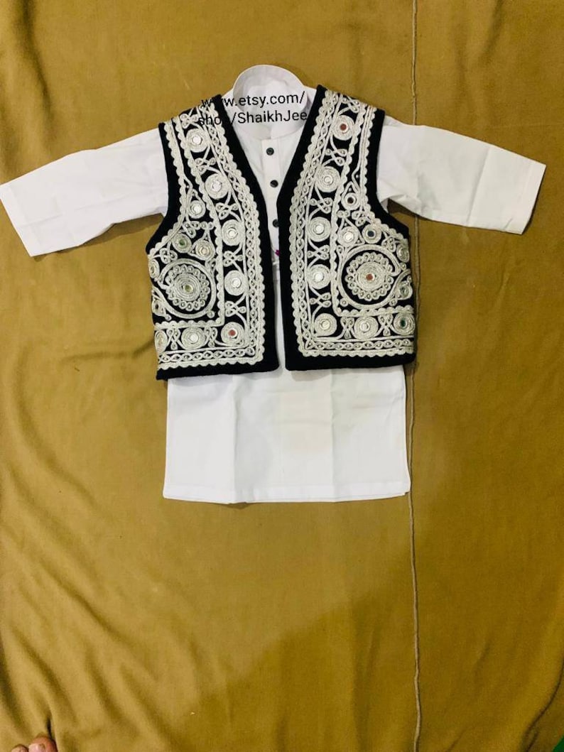 May include: A white kurta and matching pants set with a black vest. The vest is decorated with intricate silver embroidery and small reflective mirrors. The outfit is displayed on a tan surface.