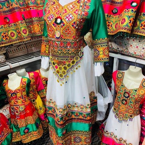 May include: A display of colourful, embroidered dresses. The dresses feature intricate patterns in red, green, gold, and white. The dresses are on mannequins, with a yellow head covering on the main dress. The dresses are likely traditional garments.
