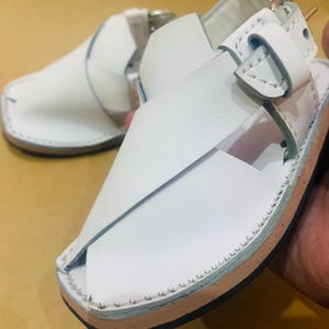 May include: White leather sandals with a square toe and cross-strap design. The sandals feature a buckle closure and a stitched sole with a light brown and blue trim. The footwear is designed for comfort and style.