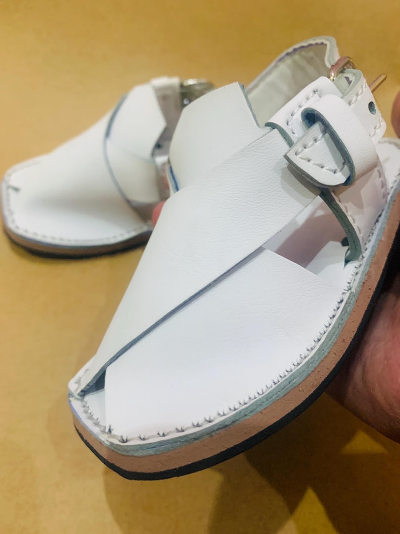 May include: White leather sandals with a square toe and cross-strap design. The sandals feature a buckle closure and a stitched sole with a light brown and blue trim. The footwear is designed for comfort and style.