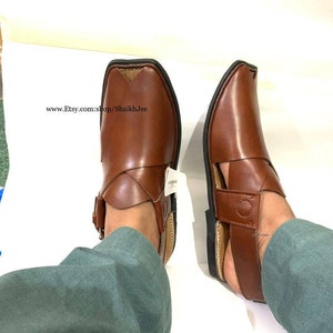 K&ouml;nnte beinhalten: Ein Paar braune Peshawari-Sandalen aus Leder mit quadratischer Spitze und Schnallenverschluss. Die Sandalen haben einen Riemen &uuml;ber dem Oberteil und eine schwarze Sohle. Der Etsy-Shop-Name ist sichtbar.