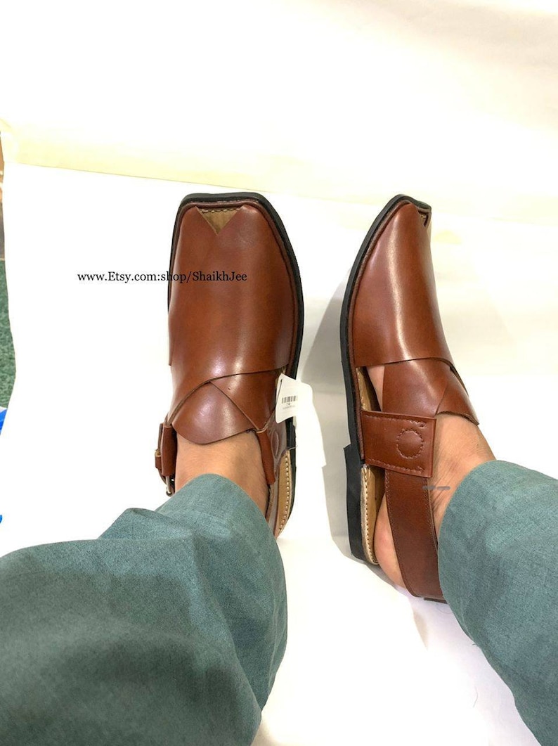 K&ouml;nnte beinhalten: Ein Paar braune Peshawari-Sandalen aus Leder mit quadratischer Spitze und Schnallenverschluss. Die Sandalen haben einen Riemen &uuml;ber dem Oberteil und eine schwarze Sohle. Der Etsy-Shop-Name ist sichtbar.
