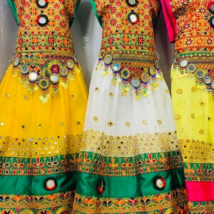 May include: Three vibrant dresses with detailed embroidery in gold, red, green, and orange. The skirts are yellow, white, and yellow. Each dress is embellished with mirrors and decorative elements. The dresses are on display.