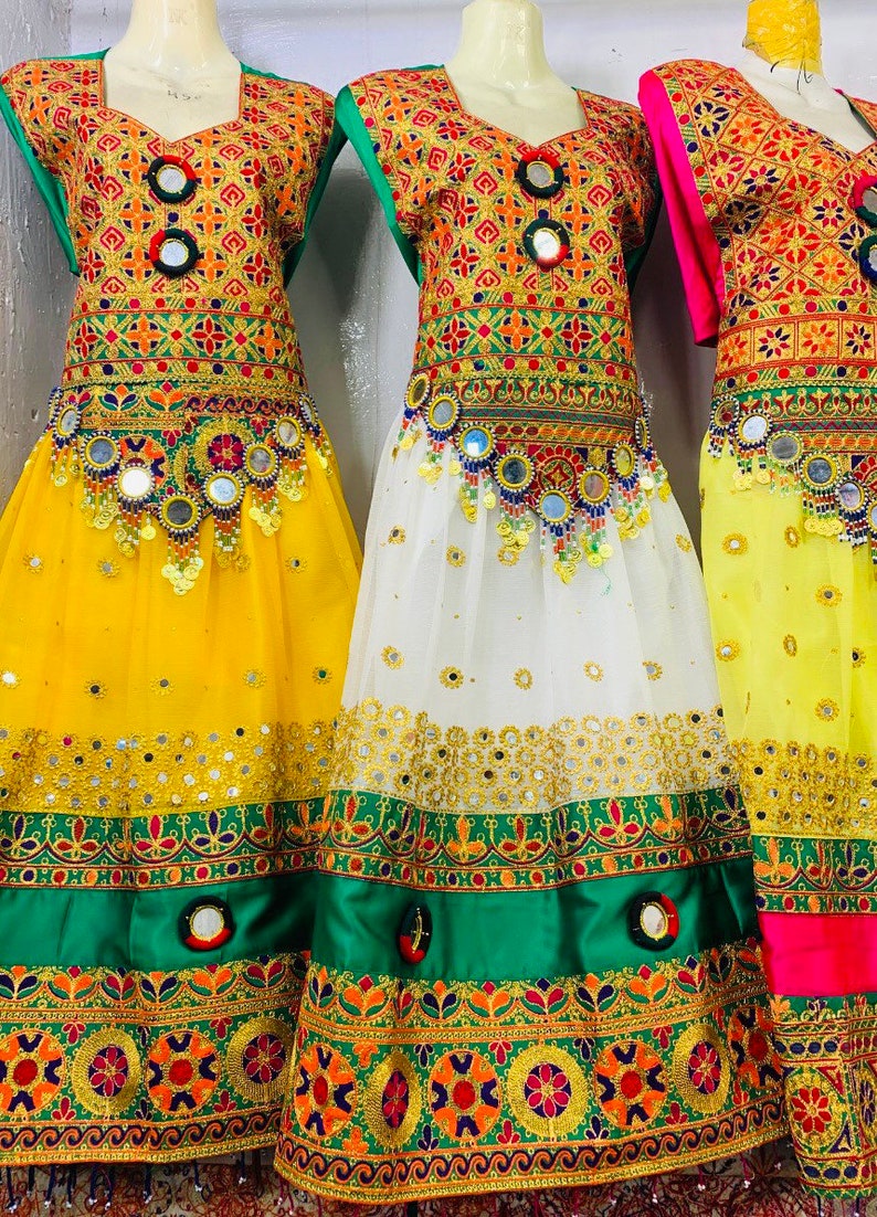 May include: Three vibrant dresses with detailed embroidery in gold, red, green, and orange. The skirts are yellow, white, and yellow. Each dress is embellished with mirrors and decorative elements. The dresses are on display.