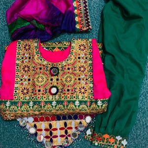 May include: A traditional outfit with a bright pink top, adorned with gold embroidery and mirrors. It's paired with green trousers and a multi-coloured scarf. The outfit displays detailed craftsmanship and cultural design.