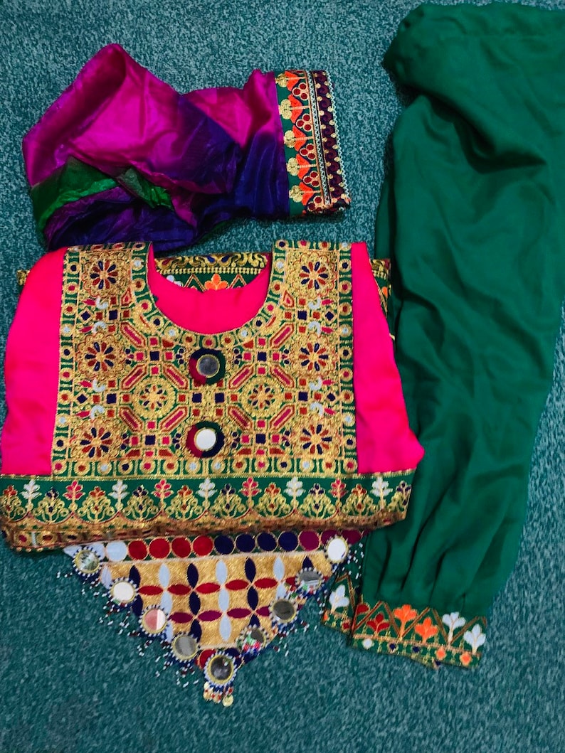 May include: A traditional outfit with a bright pink top, adorned with gold embroidery and mirrors. It's paired with green trousers and a multi-coloured scarf. The outfit displays detailed craftsmanship and cultural design.