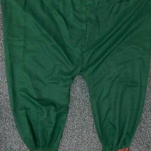 May include: Green harem trousers with embroidered cuffs. The trousers are a solid, deep green colour. The cuffs feature a detailed pattern with red, orange, blue, and gold embroidery. The trousers are made of a soft, flowing fabric.
