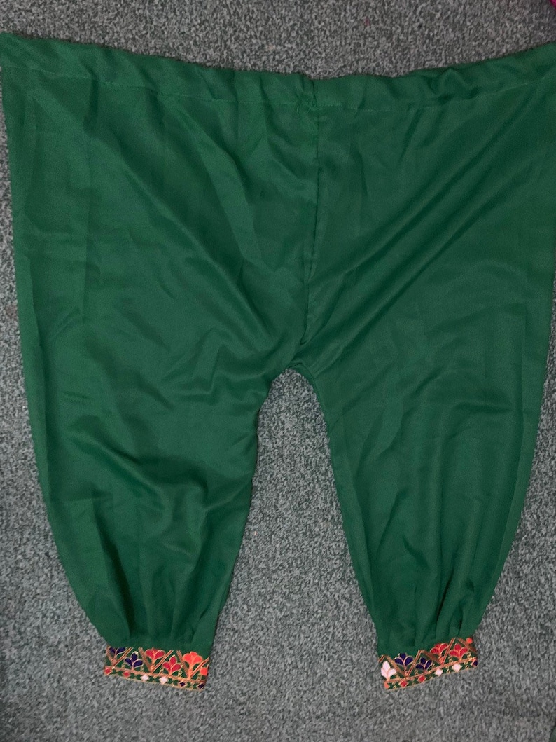 May include: Green harem trousers with embroidered cuffs. The trousers are a solid, deep green colour. The cuffs feature a detailed pattern with red, orange, blue, and gold embroidery. The trousers are made of a soft, flowing fabric.
