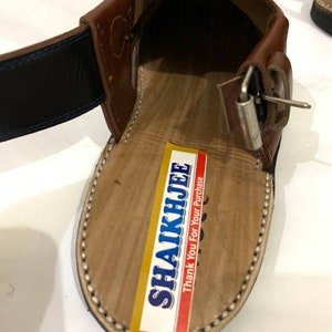 K&ouml;nnte beinhalten: Nahaufnahme einer braunen Ledersandale mit Schnallenverschluss. Die Sandale hat eine hellbraune Innensohle mit einer gen&auml;hten Bord&uuml;re. Ein Aufkleber mit dem Text "SHAIKHJEE Thank You For Your Purchase" ist sichtbar.