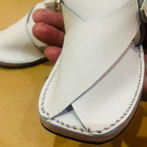May include: White leather sandals with a crisscross design over the foot. The sandals have a stitched border and a buckle closure. The sole is brown. The sandals are designed for warm weather.