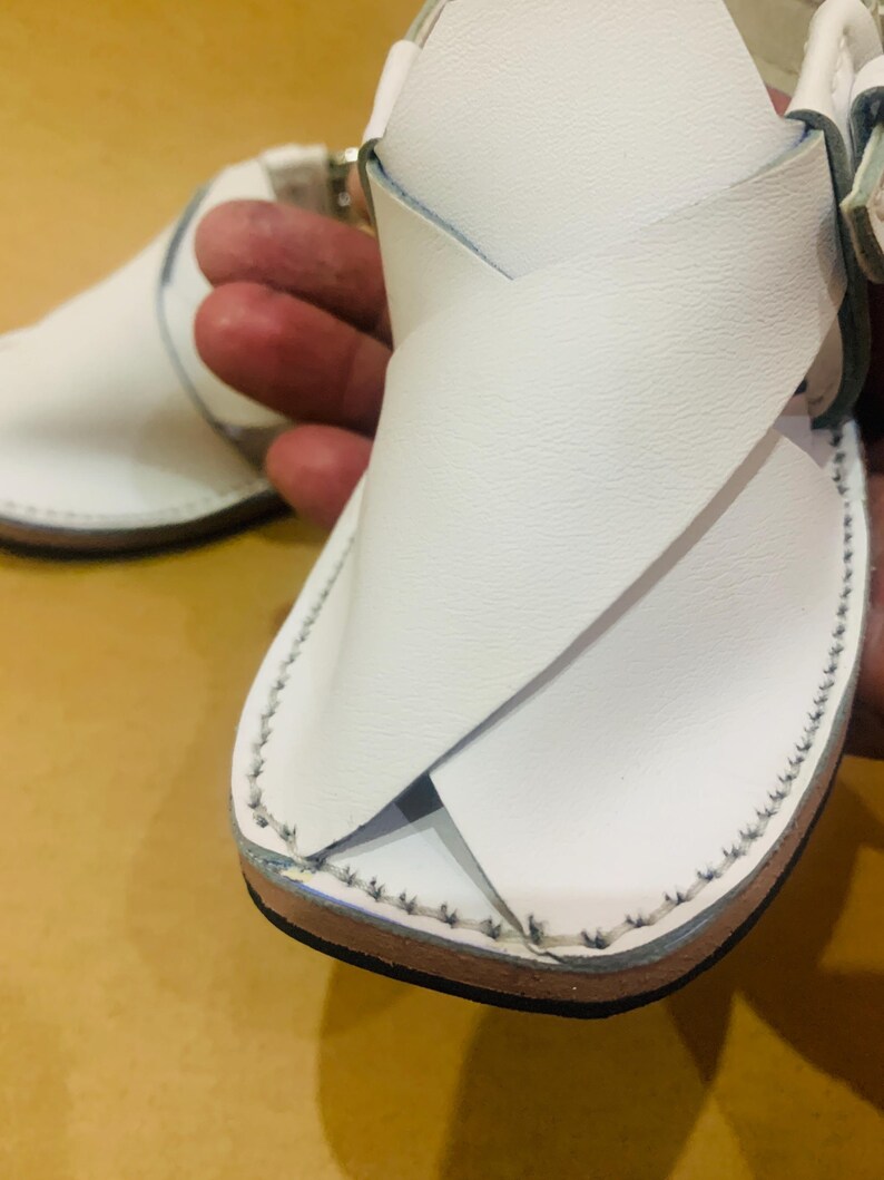 May include: White leather sandals with a crisscross design over the foot. The sandals have a stitched border and a buckle closure. The sole is brown. The sandals are designed for warm weather.