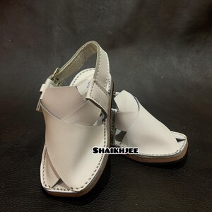 May include: A pair of white leather sandals with a strap and buckle closure. The sandals have a light brown sole and are stitched around the edges. The text "SHAIKHJEE" is visible on the side of the sandal.