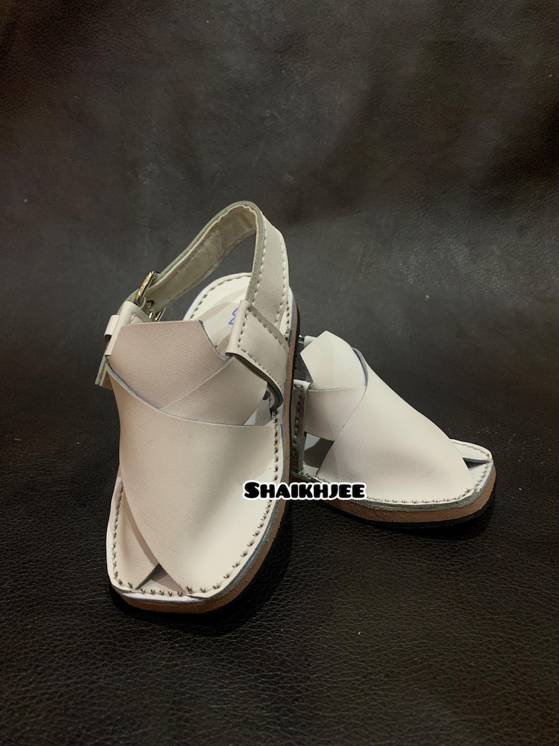 May include: A pair of white leather sandals with a strap and buckle closure. The sandals have a light brown sole and are stitched around the edges. The text "SHAIKHJEE" is visible on the side of the sandal.