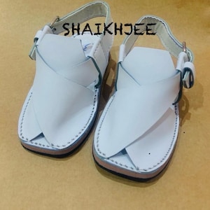 May include: A pair of white leather sandals with a cross-strap design. The sandals have a buckle closure and are stitched around the edges. The text "SHAIKHJEE" is printed on the sandals.