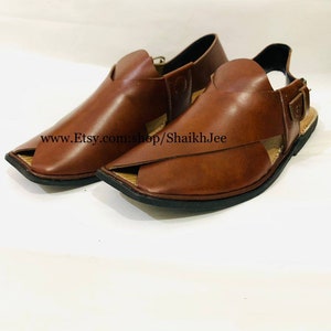 K&ouml;nnte beinhalten: Ein Paar braune Peshawari-Sandalen aus Leder. Diese Sandalen mit offener Spitze haben ein unverwechselbares Design mit einem Riemen &uuml;ber der Oberseite und einem Schnallenverschluss. Die Sohlen sind schwarz, und die Sandalen werden auf wei&szlig;em Hintergrund pr&auml;sentiert.
