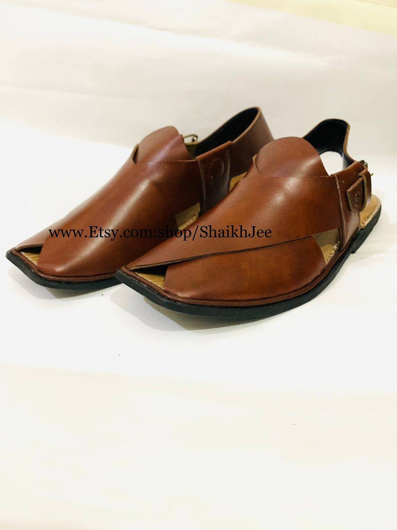 K&ouml;nnte beinhalten: Ein Paar braune Peshawari-Sandalen aus Leder. Diese Sandalen mit offener Spitze haben ein unverwechselbares Design mit einem Riemen &uuml;ber der Oberseite und einem Schnallenverschluss. Die Sohlen sind schwarz, und die Sandalen werden auf wei&szlig;em Hintergrund pr&auml;sentiert.