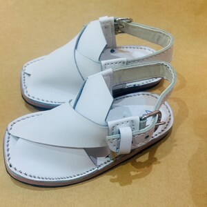 May include: A pair of white leather sandals with a strap and buckle closure. The sandals have a stitched sole and a simple design, suitable for casual wear. The sandals are displayed on a light brown surface.