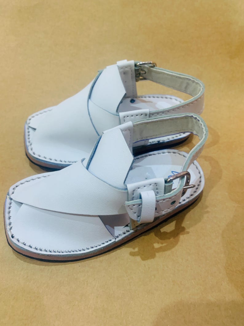May include: A pair of white leather sandals with a strap and buckle closure. The sandals have a stitched sole and a simple design, suitable for casual wear. The sandals are displayed on a light brown surface.