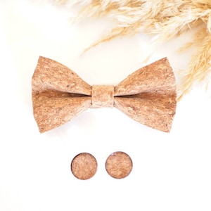 Może przedstawiać: Brązowy krawat muszkowy z korka i pasujące spinki do mankietów. Muszka wykonana jest z naturalnego korka i ma rustykalny wygląd. Spinki do mankietów są okrągłe i mają podobną teksturę korka.