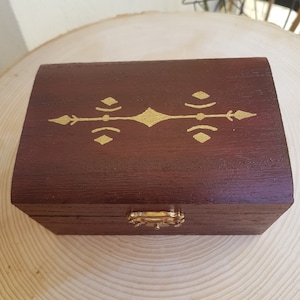 Könnte beinhalten: Eine kleine Holzkiste mit einer dunkelbraunen Beize und einem goldenen Verschluss. Die Oberseite der Kiste ist mit einem goldenen geometrischen Muster bemalt.