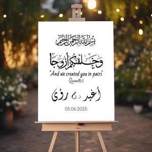 May include: A white rectangular sign on a wooden easel featuring black Arabic calligraphy, the English text "And we created you in pairs" with "Quran 78:8" below it, two names in Arabic script separated by an ampersand, and the date 05.06.2025.