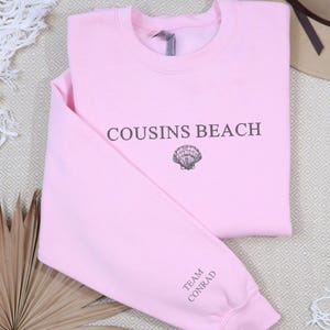 Peut inclure: Un sweat-shirt rose clair avec l'inscription "COUSINS BEACH" et un dessin de coquillage. Le texte "TEAM CONRAD" est imprimé sur la manche. Le sweat-shirt est plié sur une surface à motifs.