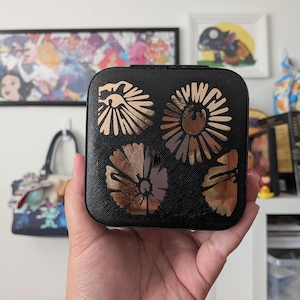 May include: A square black jewelry box with a textured surface featuring four metallic bronze floral designs on the lid. The box has rounded corners and a sleek finish, providing a compact storage solution for small accessories and personal items.