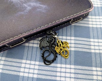 May include: A black and gold dragon charm with a silver chain. The charm is hanging from a black leather notebook.