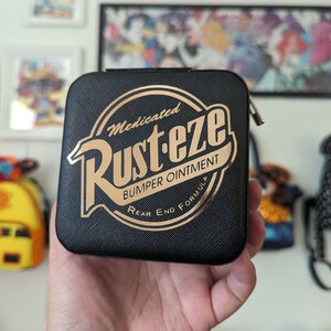 May include: A black, square-shaped, zippered pouch with a gold logo that reads "Medicated Rust-eze Bumper Ointment Rear End Formula".