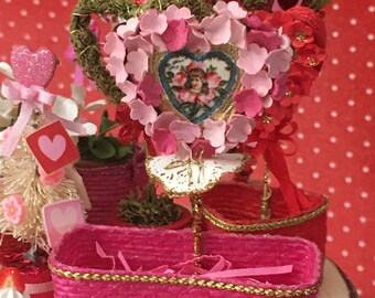 May include: A pink wicker basket with a gold trim sits on a wooden table. The basket is filled with pink and white paper hearts and a small bouquet of pink flowers. There is a small bag of colorful candies in the foreground.