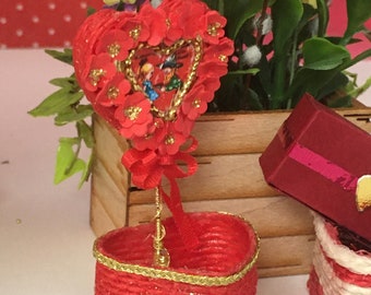 May include: A red heart-shaped basket with a gold trim, filled with a red heart-shaped floral arrangement with a gold trim. The floral arrangement has a small picture of a couple in the center.
