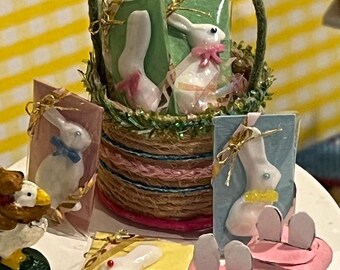May include: A basket filled with wrapped Easter bunny shaped treats, along with other bunny shaped treats on a white surface. The basket is made of woven twine and has a green handle. The treats are wrapped in clear plastic and have a white bunny with pink ears and a pink bow. The other treats are on a yellow, pink, and blue background.