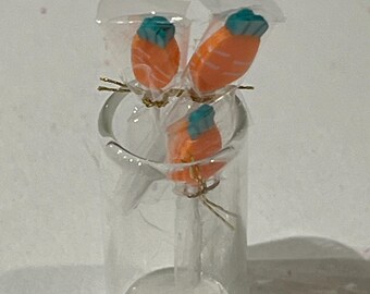 May include: Three orange and teal carrot-shaped lollipops are displayed in a clear glass jar. The lollipops are wrapped in clear plastic.