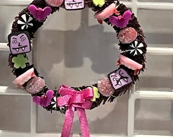 May include: A brown Halloween wreath decorated with pink, purple, and green candy, bats, and vampire faces. A pink bow hangs from the bottom of the wreath.
