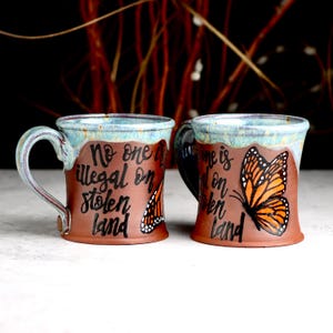 May include: Two ceramic mugs with brown bodies and blue-green rims. Each mug features the text "No one is illegal on stolen land" and a monarch butterfly illustration. The mugs have curved handles.
